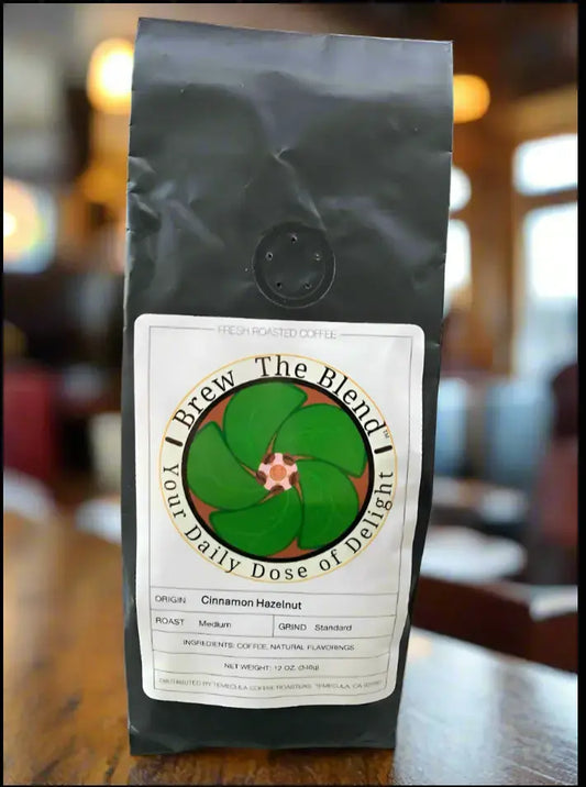 A bag of Brew The Blend Cinnamon Hazelnut coffee sits on a wooden table with a blurred cafe background. The bag is matte black with a white label featuring a green flower logo.